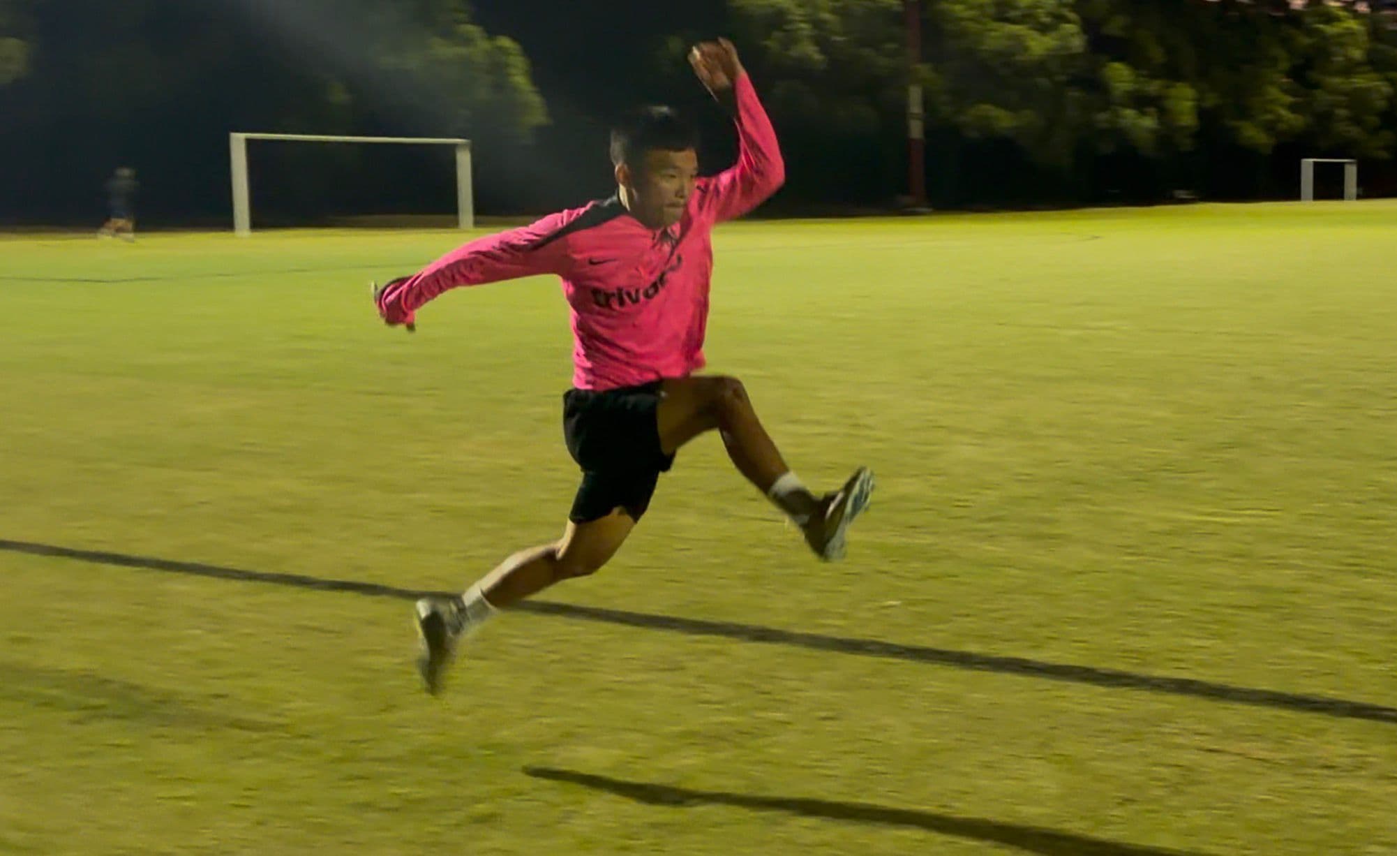 Athlete mid-flight in a horizontal bound under stadium lights — knee driven up, opposite arm in counter-balance, elastic energy releasing into horizontal projection