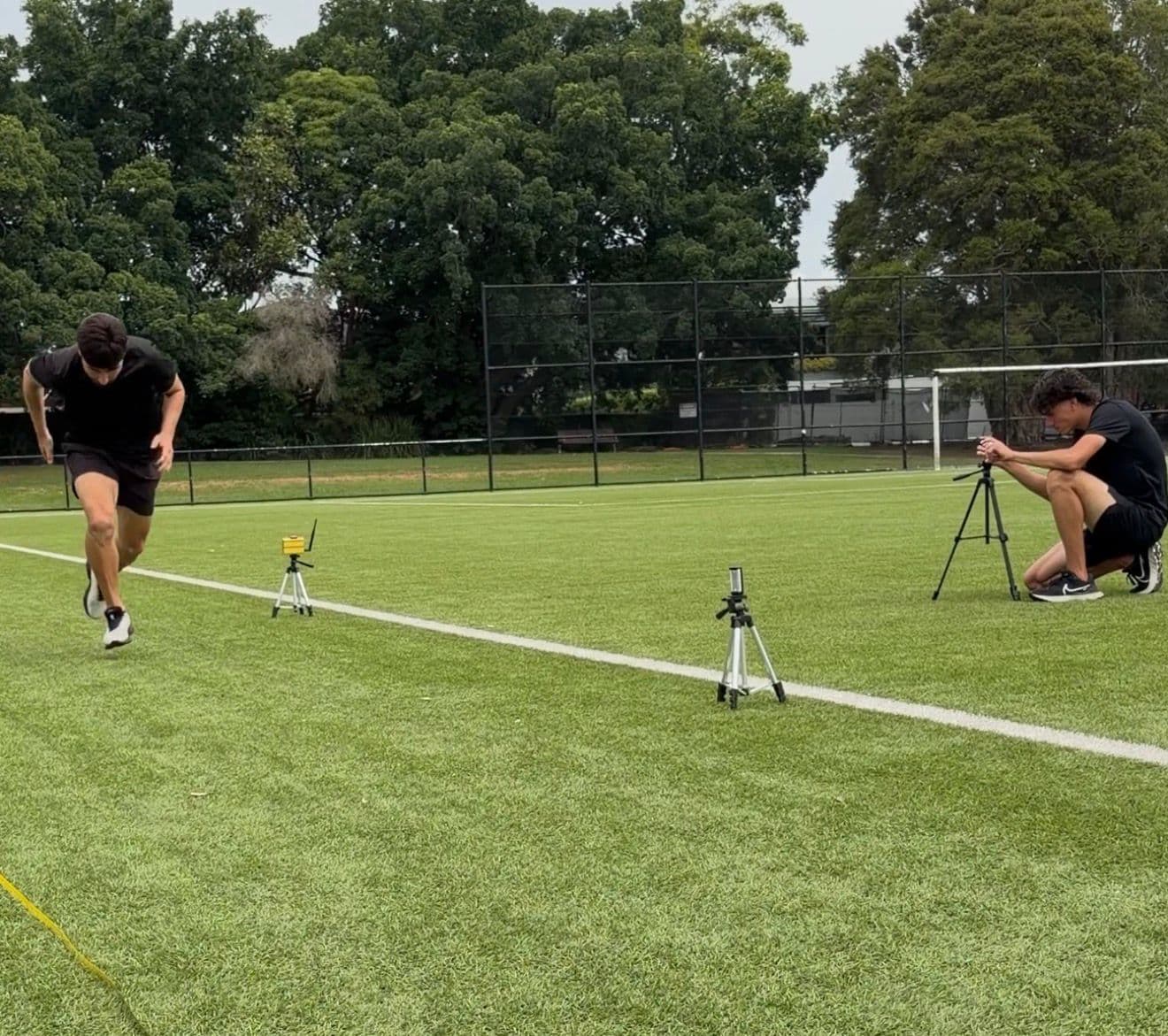 Athlete accelerating past electronic timing gates while a coach films from a tripod